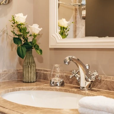 Ein Waschbecken mit einer Marmorablage, einer glänzenden Armatur und einem zusammengelegten weißen Handtuch. Daneben steht ein Trinkglas und eine Vase mit weißen Rosen. Ein Spiegel mit weißem Rahmen hängt über dem Waschbecken.