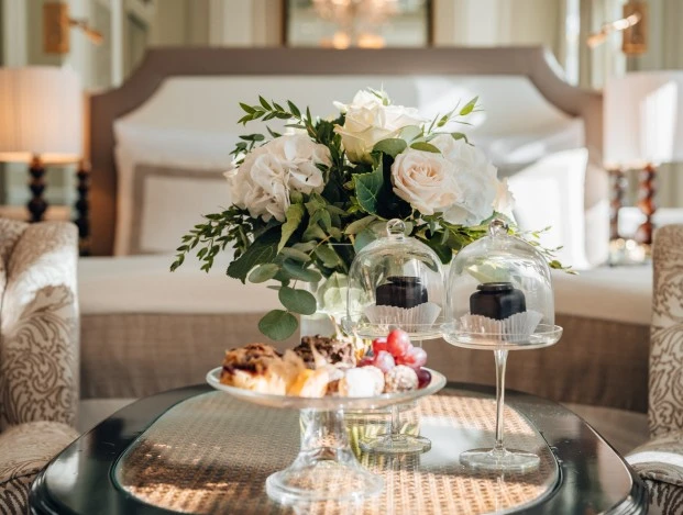 Auf dem Tisch stehen zwei Original Sacher Würfel unter einer Glashaube. Daneben befindet sich ein Blumenstrauß mit weißen Rosen und Hortensien. Im Hintergrund ist ein Bett mit weißen Kissen und einer gepolsterten Kopflehne zu sehen. Rechts und links neben dem Bett stehen Nachttischlampen.