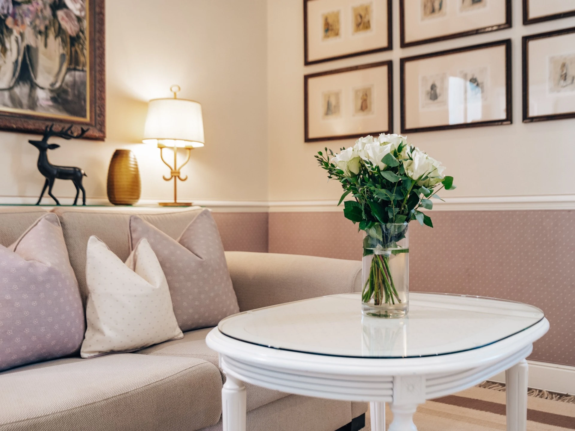 Gemütliche Sitzecke mit beigem Sofa, pastellfarbenen Kissen und einem weißen Couchtisch, auf dem ein frischer Blumenstrauß mit weißen Rosen steht. Dezente Wandgestaltung mit gerahmten Illustrationen und eine warme Beleuchtung schaffen eine behagliche Atmosphäre.