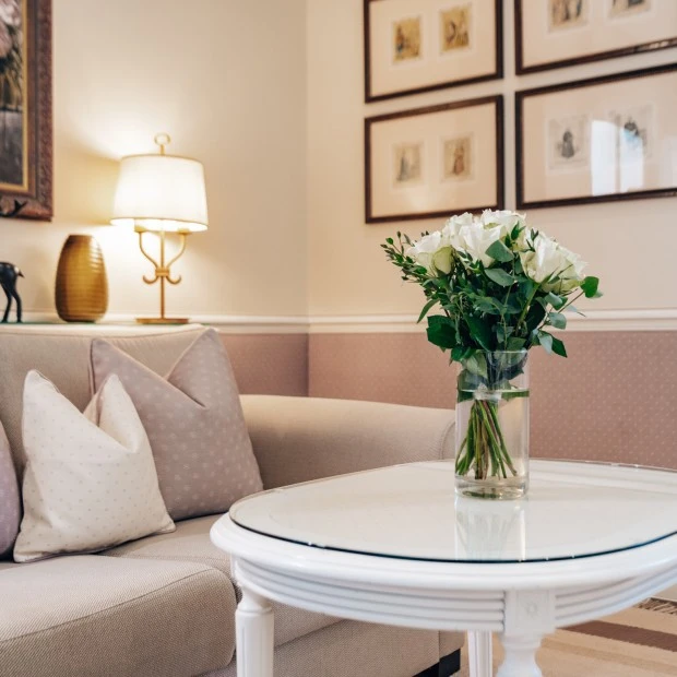 Gemütliche Sitzecke mit beigem Sofa, pastellfarbenen Kissen und einem weißen Couchtisch, auf dem ein frischer Blumenstrauß mit weißen Rosen steht. Dezente Wandgestaltung mit gerahmten Illustrationen und eine warme Beleuchtung schaffen eine behagliche Atmosphäre.