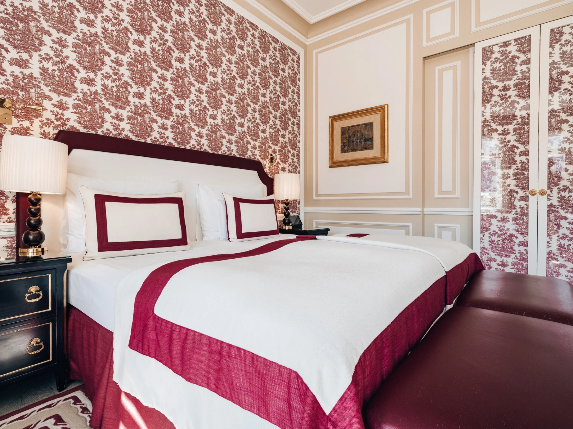Detailansicht eines klassisch eingerichteten Hotelzimmers mit rot-weißer Bettwäsche, gemusterter Tapete in Bordeauxrot und beiger Wandvertäfelung, ergänzt durch ein stimmungsvolles Wandbild und eine stilvolle Nachttischlampe.