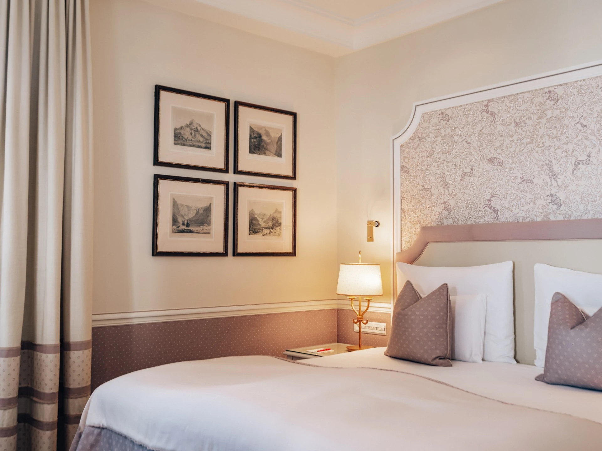 Gemütliches Hotelzimmer mit einem großen Doppelbett, verziert mit dekorativen Kissen und einem floralen Stoffpaneel als Kopfteil. An der Wand hängen vier gerahmte historische Landschaftszeichnungen. Warme Beleuchtung durch eine klassische Wandlampe neben dem Bett sorgt für eine einladende Atmosphäre. Helle Vorhänge rahmen das Fenster sanft ein.