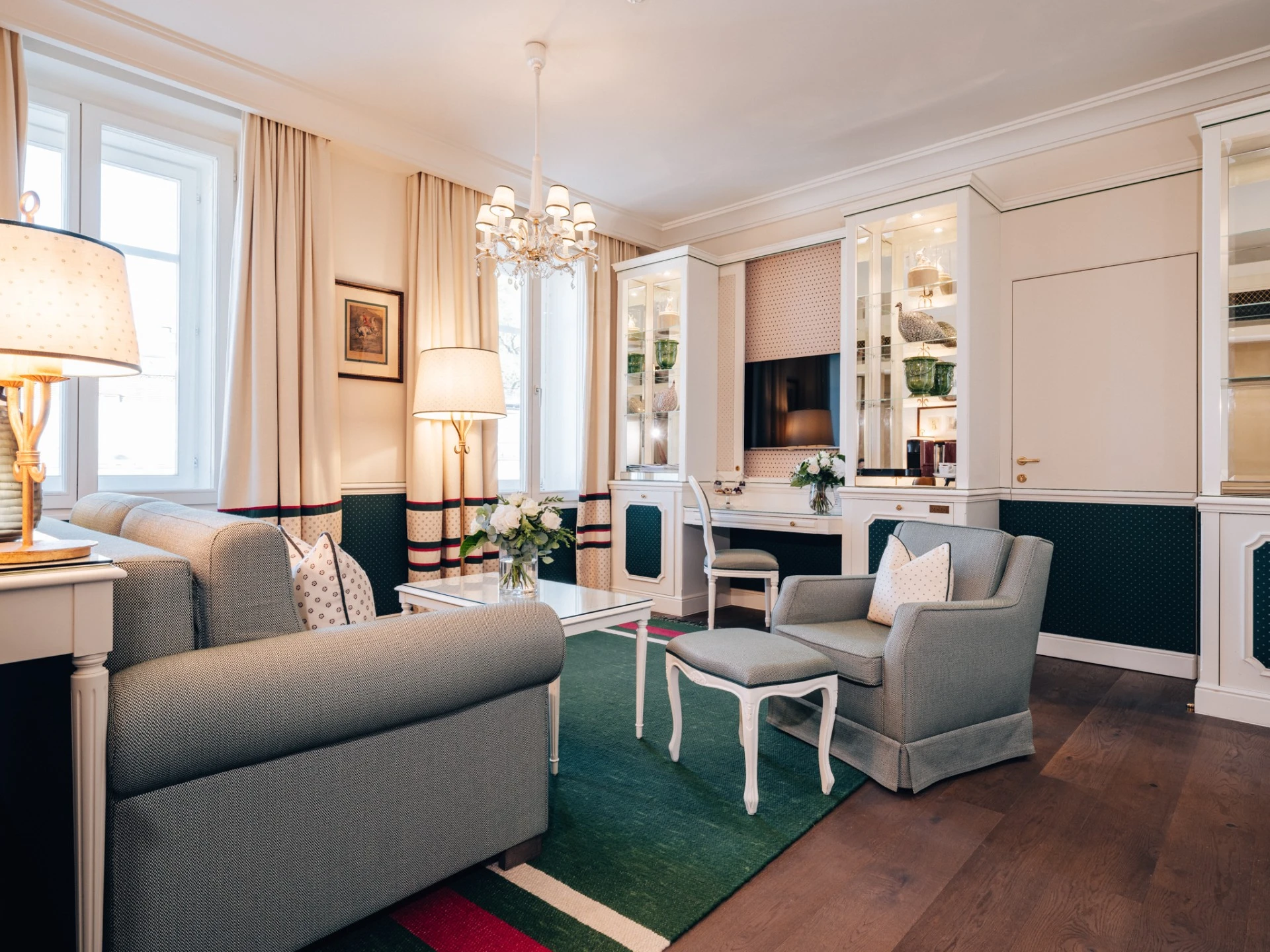 Heller, eleganter Wohnbereich mit klassischem Interieur in Grün-, Creme- und Rottönen. Zwei Sofas, ein Sessel mit Hocker und ein Couchtisch mit frischen Blumen sorgen für Gemütlichkeit. Im Hintergrund integrierte Vitrinen mit Kristall und Porzellan sowie ein Schreibtisch mit moderner Leselampe. Der Raum wirkt durch große Fenster, helle Vorhänge und stilvolle Beleuchtung offen und einladend.
