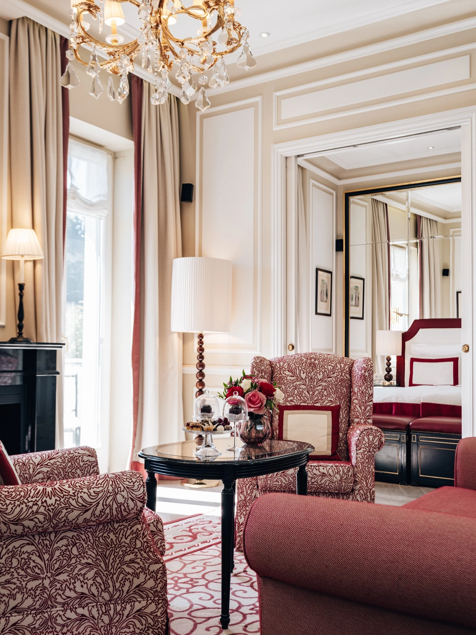 Elegantes Wohnzimmer in klassischen Rottönen mit floralen Mustern. Zwei gemütliche Sessel und ein Sofa gruppieren sich um einen schwarzen Couchtisch, auf dem frische Blumen, und Original Sacher-Würfel unter Glashauben serviert werden. Im Hintergrund der Blick in ein stilvolles Schlafzimmer mit rotem Betthaupt. Ein goldener Kronleuchter und hohe Fenster sorgen für eine helle, luxuriöse Atmosphäre.