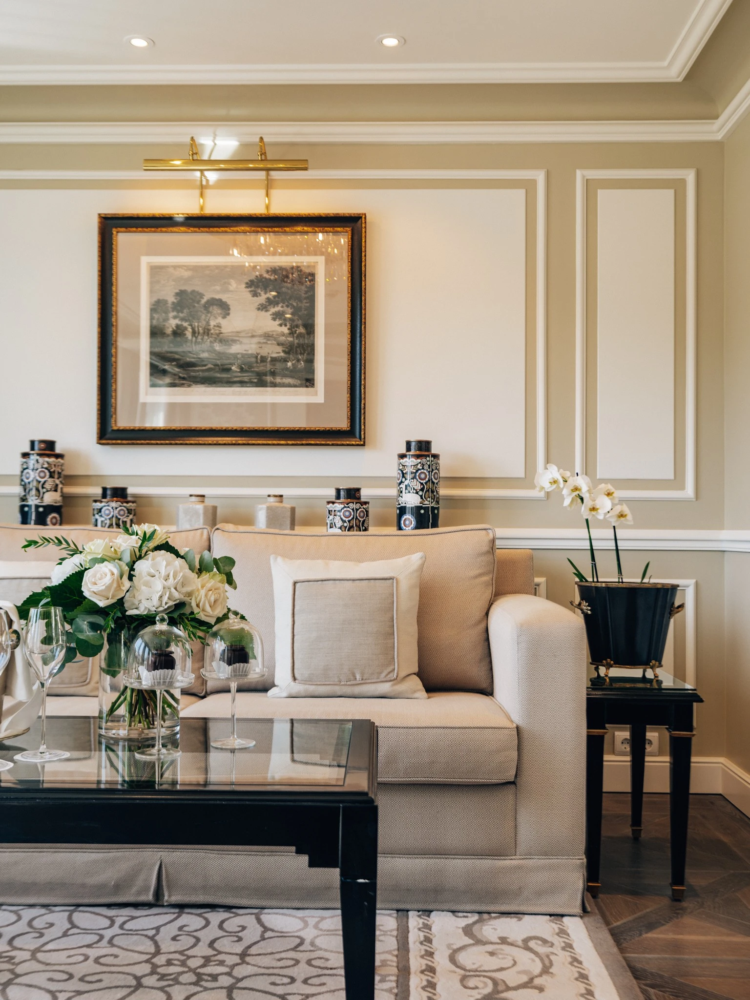 Stilvoll eingerichtete Sitzecke mit beiger Couch, floralem Blumengesteck und Original Sacher-Würfel unter Glashauben auf einem Glastisch, dekoriert mit klassischer Wandkunst und schwarzen Vasen auf Konsolen im Hintergrund.