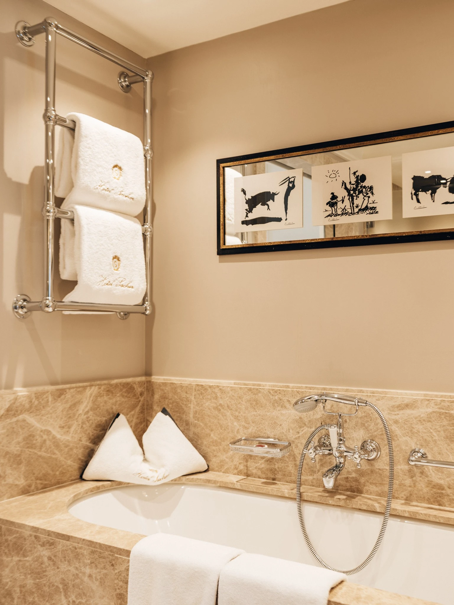 Badezimmer mit eleganter Badewanne, umrahmt von beigem Marmor. Darüber ein verchromter Handtuchhalter mit zwei flauschigen, weißen Hotelhandtüchern. An der Wand ein gerahmtes Kunstwerk mit drei minimalistischen Illustrationen. Die Armaturen im Retro-Stil und zwei Sacher-Kissen sorgen für ein stilvolles Ambiente.