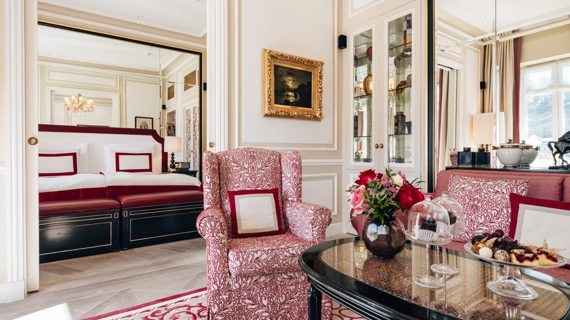 Stilvoll eingerichteter Wohnbereich einer Suite mit rot-gemusterten Polstermöbeln, Glasvitrine, frischen Blumen auf dem Couchtisch und einer elegante Etagere mit kleinen Süßigkeiten und zwei Original Sacher-Würfel unter Glashaubensowie. Blick geht auf das angrenzende Schlafzimmer mit edlem Doppelbett und eleganten Spiegel- und Stuckelementen.