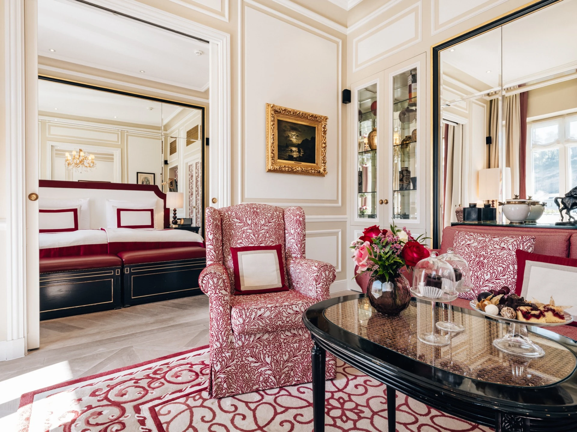 Stilvoll eingerichteter Wohnbereich einer Suite mit rot-gemusterten Polstermöbeln, Glasvitrine, frischen Blumen auf dem Couchtisch und einer elegante Etagere mit kleinen Süßigkeiten und zwei Original Sacher-Würfel unter Glashaubensowie. Blick geht auf das angrenzende Schlafzimmer mit edlem Doppelbett und eleganten Spiegel- und Stuckelementen.