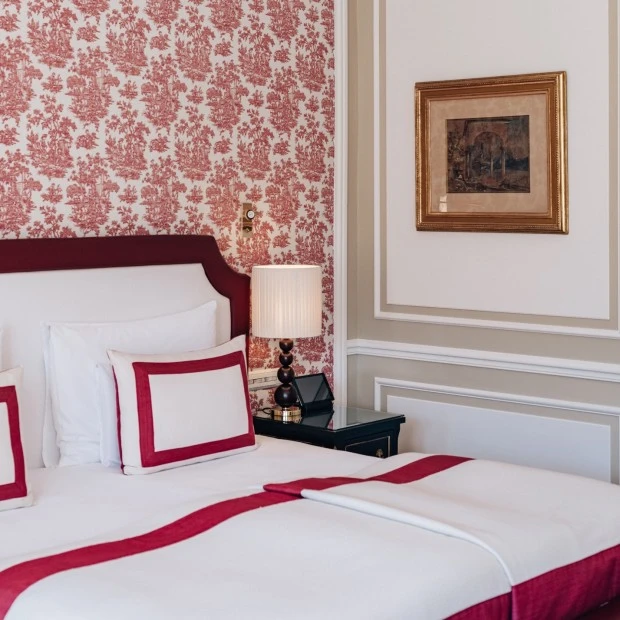 Detailansicht eines klassisch eingerichteten Hotelzimmers mit rot-weißer Bettwäsche, gemusterter Tapete in Bordeauxrot und beiger Wandvertäfelung, ergänzt durch ein stimmungsvolles Wandbild und eine stilvolle Nachttischlampe.