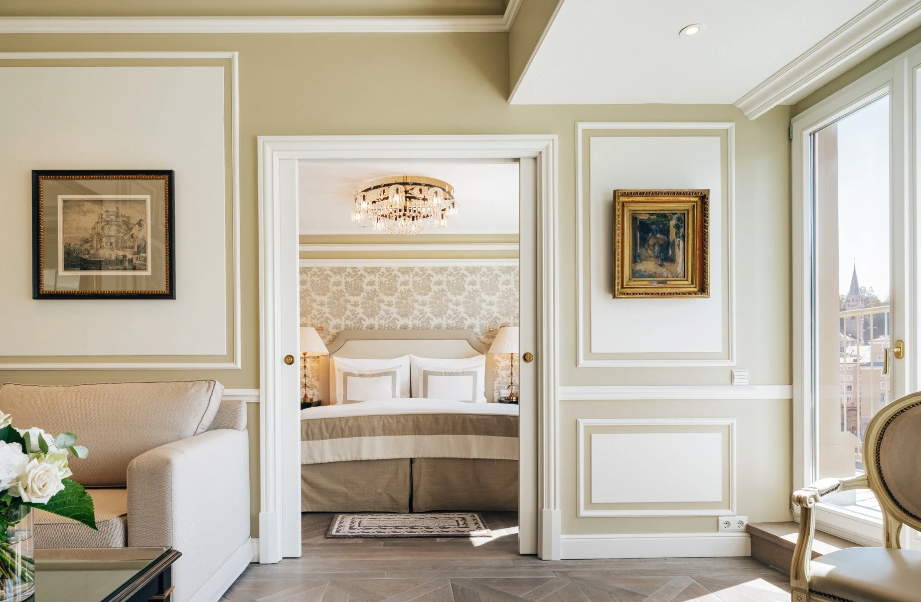 Blick vom eleganten Wohnbereich in ein klassisch eingerichtetes Schlafzimmer mit cremefarbenem Dekor, floraler Tapete und prunkvollem Kronleuchter, eingerahmt von offenen Flügeltüren.