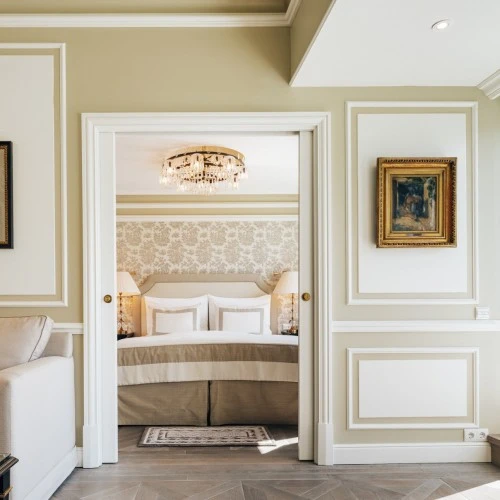 Blick vom eleganten Wohnbereich in ein klassisch eingerichtetes Schlafzimmer mit cremefarbenem Dekor, floraler Tapete und prunkvollem Kronleuchter, eingerahmt von offenen Flügeltüren.