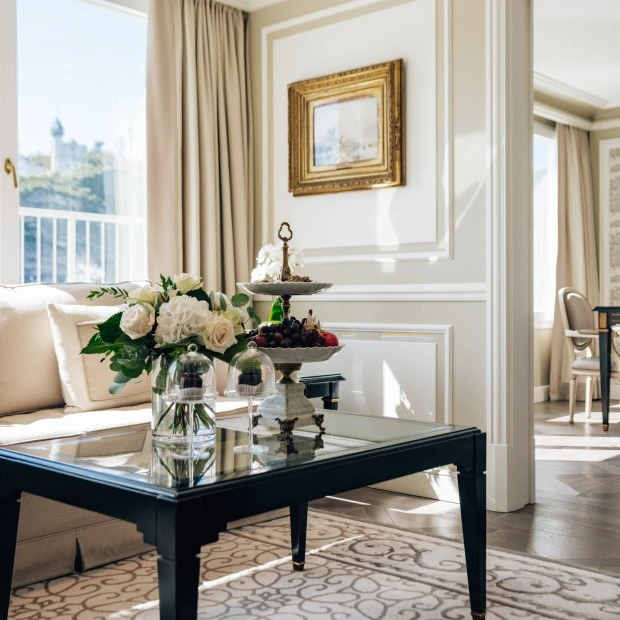 Lichtdurchflutetes, elegantes Wohnzimmer mit beiger Couch und Blick in ein weiteres Zimmer mit Tisch und Stühlen. Auf dem Beistelltisch vor der Couch stehen ein dekoratives Blumengesteck und daneben eine elegante Etagere mit frischen Früchten und Original Sacher-Würfel unter Glashauben.