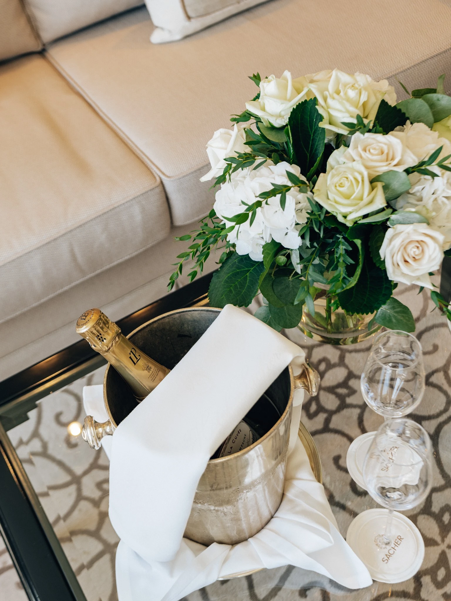 Champagnerkühler mit Flasche und weißen Stoffservietten, daneben ein eleganter Blumenstrauß aus weißen Rosen und Hortensien auf einem Glastisch im stilvoll eingerichteten Hotelzimmer.