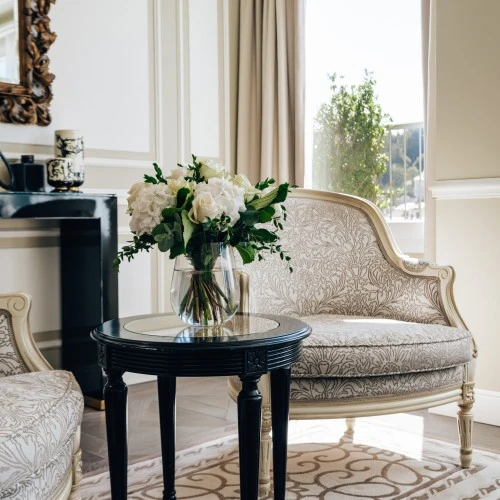 Zwei elegante, beige-gemusterte Sessel um einen kleinen runden Tisch mit Glasvase und frischem Blumenstrauß, im Hintergrund ein großes Fenster wo die Sonne rein strahlt