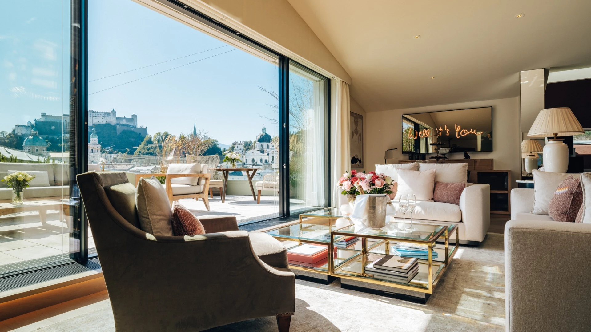 Lichtdurchflutetes Wohnzimmer der Sky Suite mit eleganter Sitzecke, pastellfarbenen Kissen, frischen Blumenarrangements und Champagnerkühler auf dem Couchtisch und direktem Zugang zur großzügigen Terrasse mit Blick über die Stadt Salzburg.