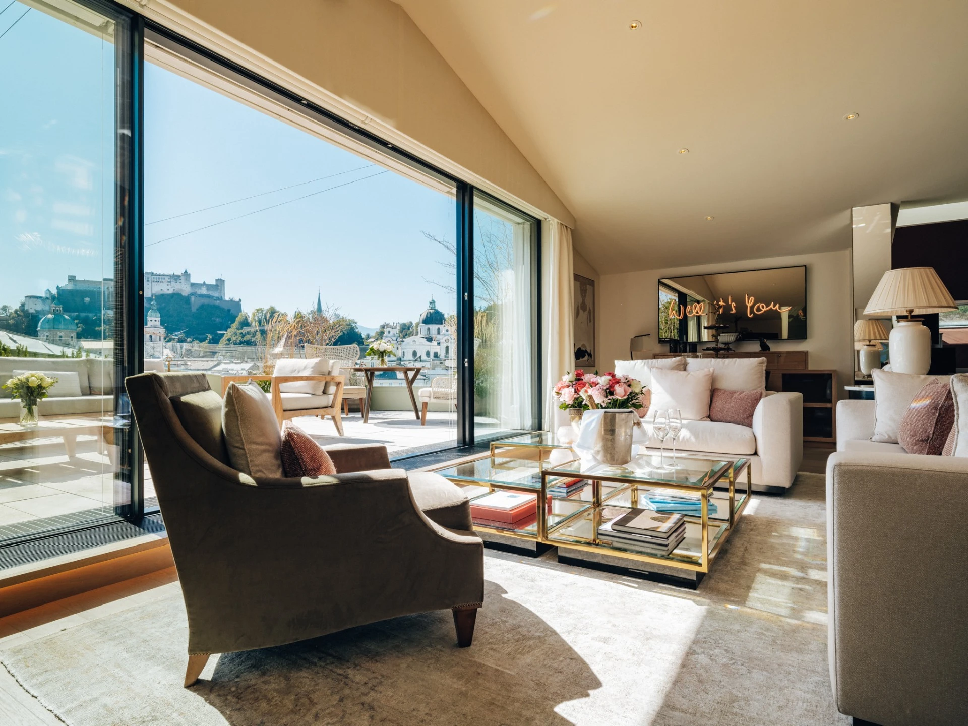 Lichtdurchflutetes Wohnzimmer der Sky Suite mit eleganter Sitzecke, pastellfarbenen Kissen, frischen Blumenarrangements und Champagnerkühler auf dem Couchtisch und direktem Zugang zur großzügigen Terrasse mit Blick über die Stadt Salzburg.