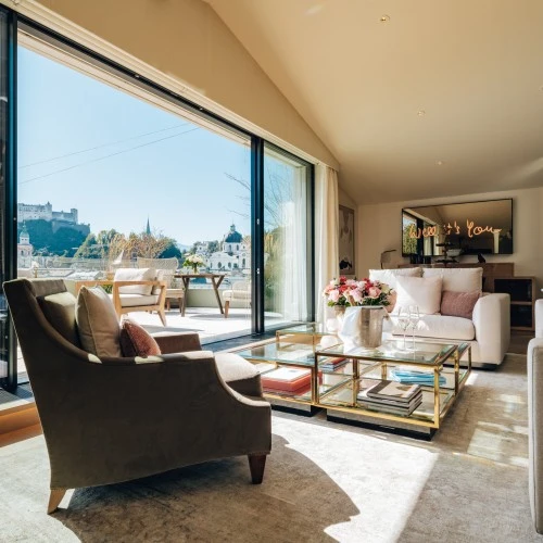 Lichtdurchflutetes Wohnzimmer der Sky Suite mit eleganter Sitzecke, pastellfarbenen Kissen, frischen Blumenarrangements und Champagnerkühler auf dem Couchtisch und direktem Zugang zur großzügigen Terrasse mit Blick über die Stadt Salzburg.