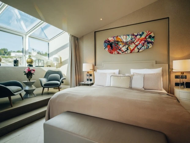 Sky-Suite_HSS_092023_Web_01 (c) Hotel Sacher Elegantes Schlafzimmer mit einem großen, gemütlich Bett, modernem abstrakten Kunstwerk über dem Kopfteil, großer Fensterfront mit Blick auf die Festung Hohensalzburg. Zwei stilvolle Sessel mit Beistelltisch und frischen Blumen sorgen für eine einladende Sitzecke.
