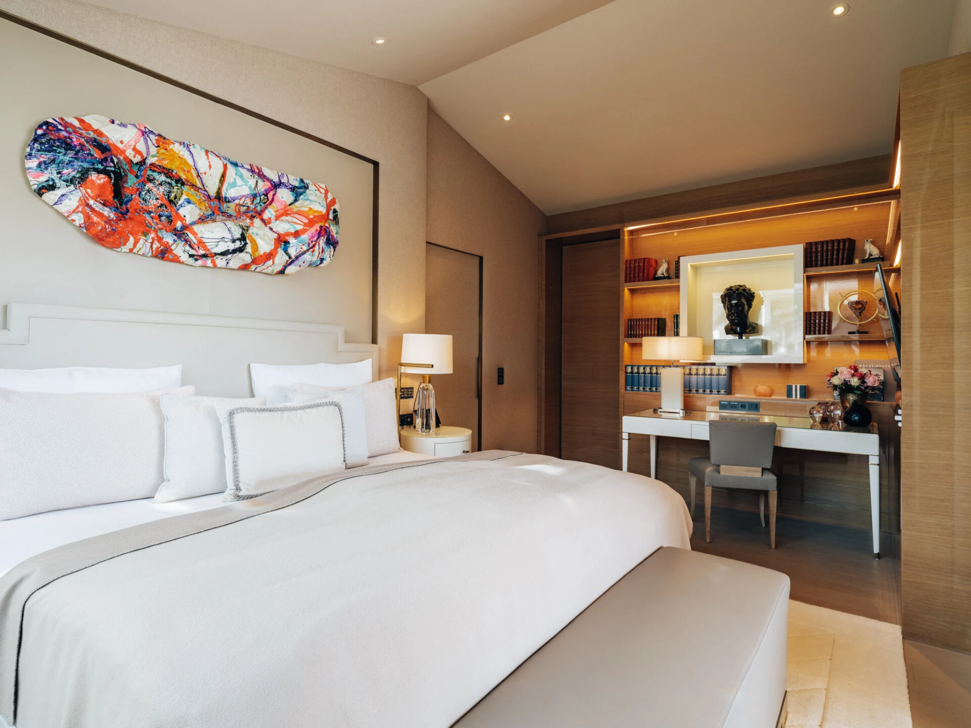 Elegantes Schlafzimmer mit einem großen, gemütlich Bett, modernem abstrakten Kunstwerk über dem Kopfteil, einem Schreibtisch mit Bücherregal, stilvoller Beleuchtung und dekorativen Elementen wie einer Skulptur und frischen Blumen.