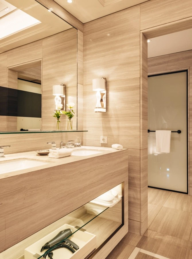 Sky-Suite_HSS_092023_Web_07 (c) Hotel Sacher Modernes, helles Badezimmer mit Doppelwaschbecken, großen Spiegeln, stilvollen Leuchten und frischen weißen Rosen; im Hintergrund ein abgetrennter Duschbereich und ein bereitgelegter Bademantel.