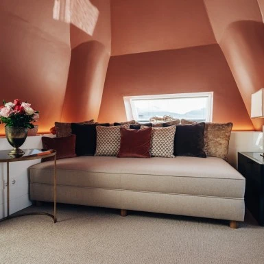 Gemütliche Ecke in der Sky Suite mit beigefarbener Recamiere, dekorativen Kissen in warmen Tönen, frischem Blumenarrangement und stilvoller Tischlampe, eingerahmt von den charakteristischen Dachschrägen und einem kleinen Fenster mit Ausblick.