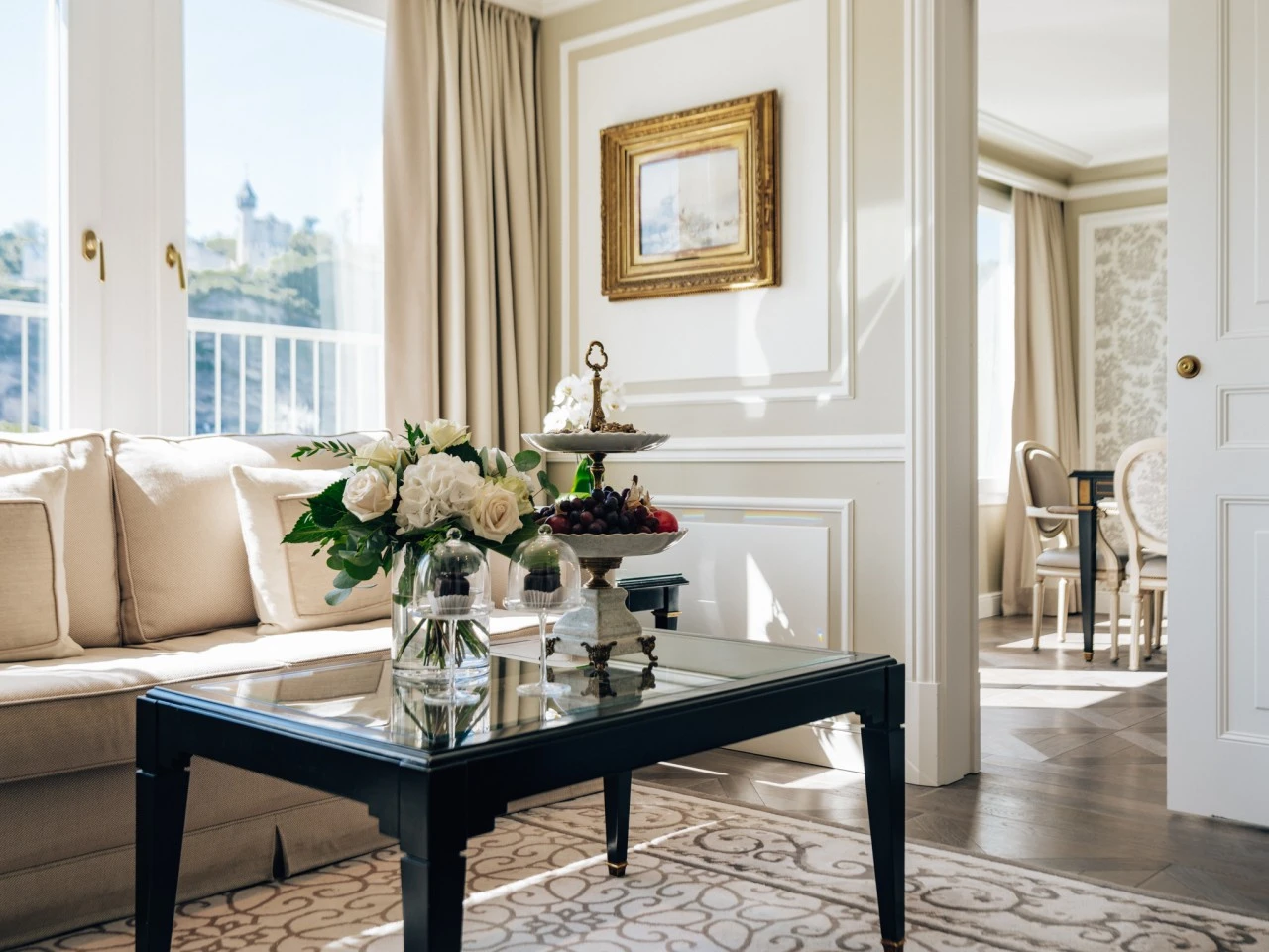 Lichtdurchflutetes Hotelzimmer mit einem stilvoll gedeckten Beistelltisch mit frischen weißen Rosen und Hortensien in einer Vase sowie einer Etagere mit kleinen Desserts und diversen Obstsorten. Dahinter der Ausblick auf den Mönchsberg.