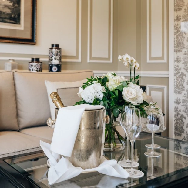 Champagnerkühler mit Flasche und weißen Stoffservietten, daneben ein eleganter Blumenstrauß aus weißen Rosen und Hortensien und zwei Etageren mit zwei Original Sacher-Würfel auf einem Glastisch im stilvoll eingerichteten Hotelzimmer.