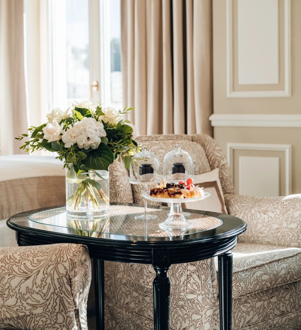 Stilvoll gedeckter Beistelltisch mit frischen weißen Rosen und Hortensien in einer Vase sowie einer Etagere mit kleinen Desserts, eingerahmt von zwei eleganten Polstersesseln vor bodenlangen Vorhängen.