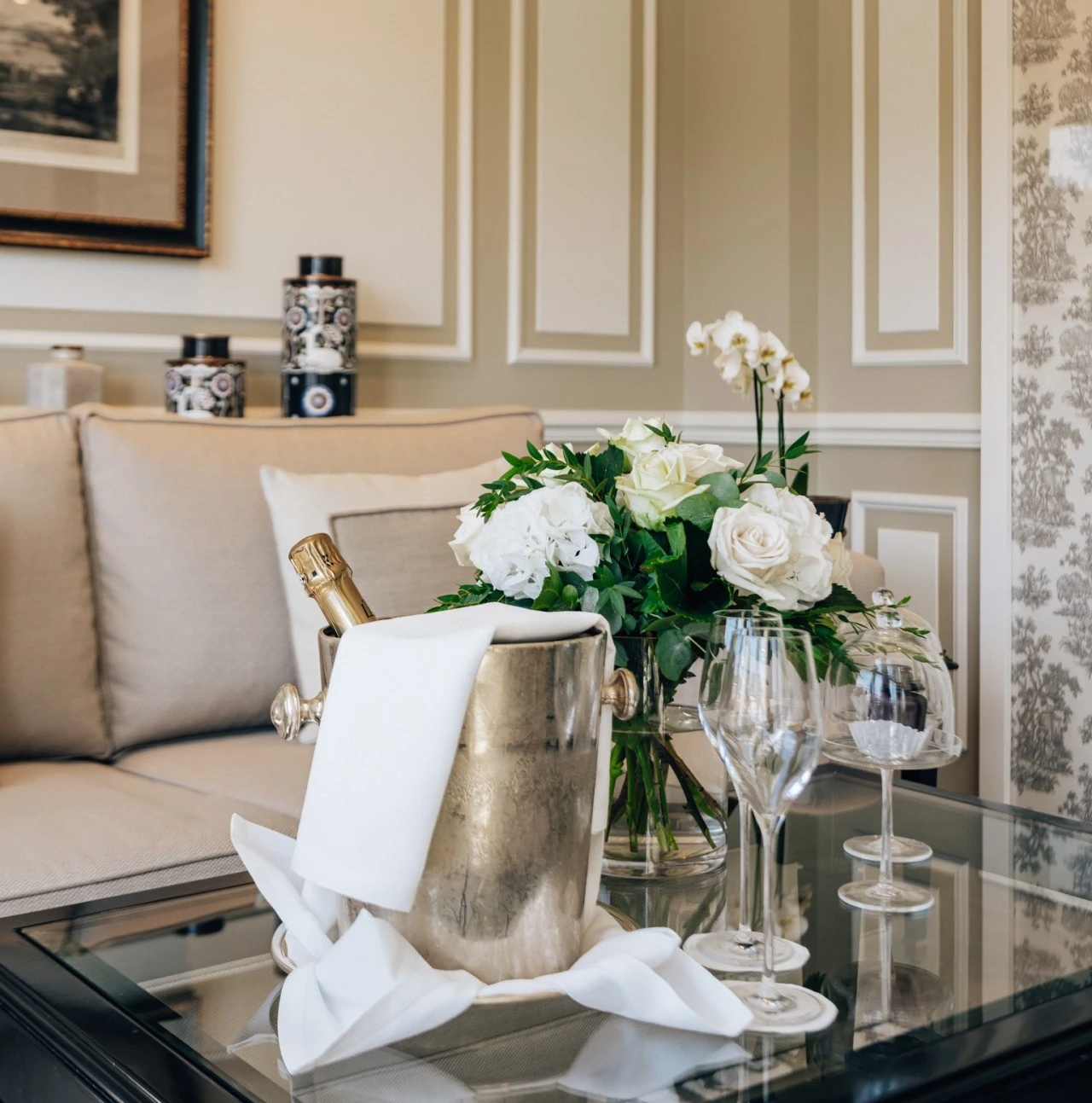 Champagnerkühler mit Flasche und weißen Stoffservietten, daneben ein eleganter Blumenstrauß aus weißen Rosen und Hortensien und zwei Etageren mit zwei Original Sacher-Würfel auf einem Glastisch im stilvoll eingerichteten Hotelzimmer.