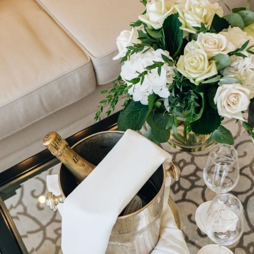 Champagnerkühler mit Flasche und weißen Stoffservietten, daneben ein eleganter Blumenstrauß aus weißen Rosen und Hortensien auf einem Glastisch im stilvoll eingerichteten Hotelzimmer.