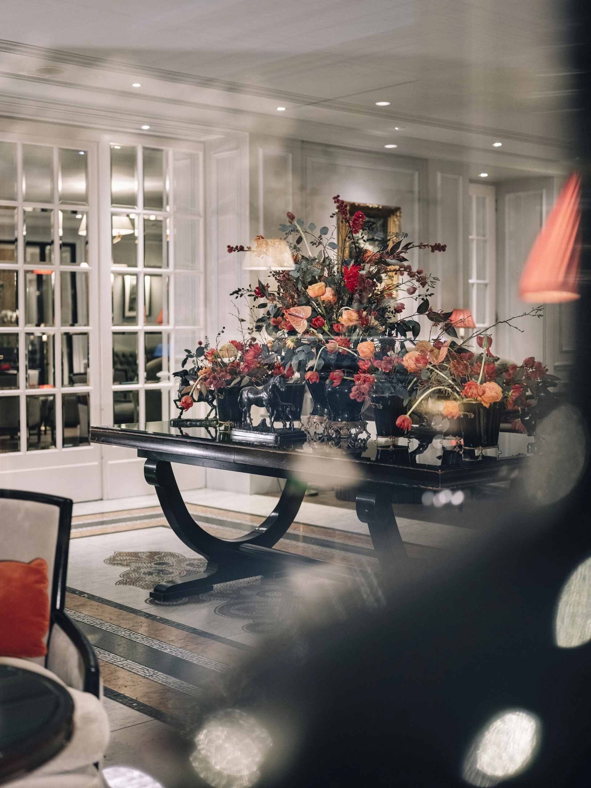 Üppiges Blumenarrangement mit roten, orangen und rosafarbenen Blüten auf einem dunklen Holztisch im eleganten, lichtdurchfluteten Empfangsbereich des Hotel Sacher Salzburg.