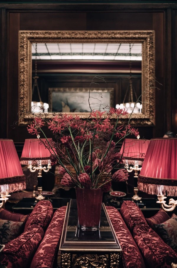Ein üppiger Blumenstrauß in kräftigen Rottönen steht auf einem edlen Tisch zwischen vier roten, reich verzierten Lampen mit Fransen, eingebettet in einen klassisch eingerichteten Salon mit dunkler Holzvertäfelung und großem Spiegel.