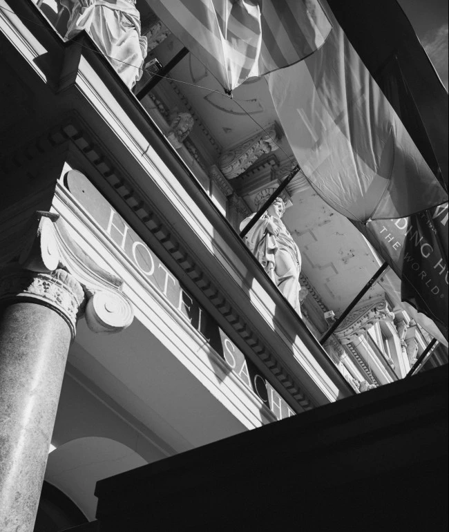 Schwarz-weiß-Aufnahme der Fassade des Hotel Sacher mit Säulen, Statuen und wehenden Fahnen im Gegenlicht.