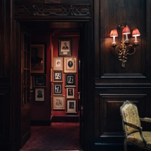 Blick in den Künstlergang des Hotel Sacher Wien mit historischen Porträts an den Wänden, dunkler Holzvertäfelung, rotem Teppich und warmem Licht von Wandlampen.