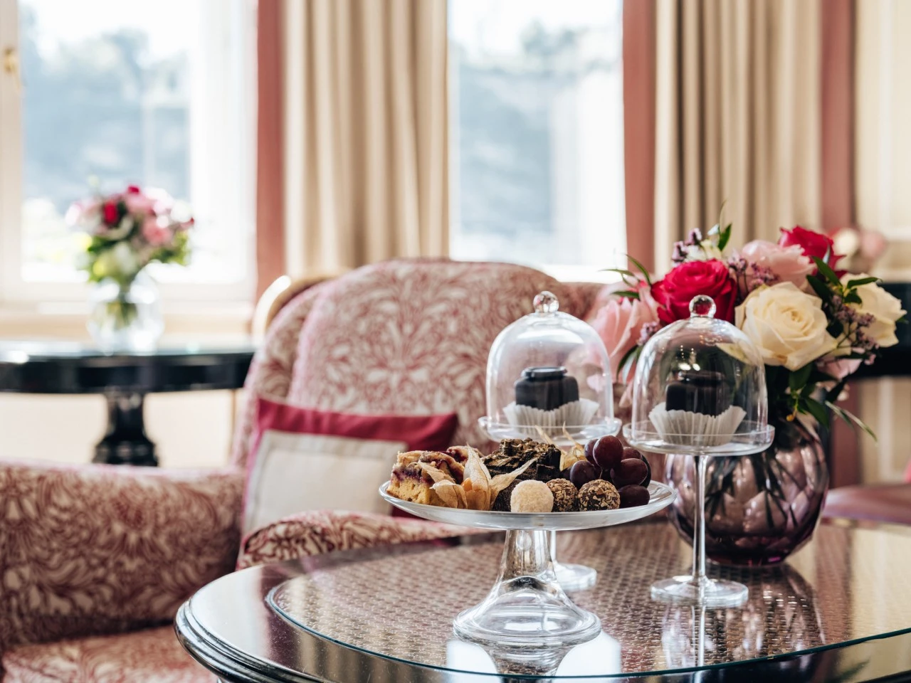 Ein Arrangement aus Pralinen, Gebäck und Original Sacher-Würfeln auf einer Etagere, umgeben von frischen Blumen, steht auf einem eleganten Glastisch in einem gemütlichen, sonnigen Zimmer.