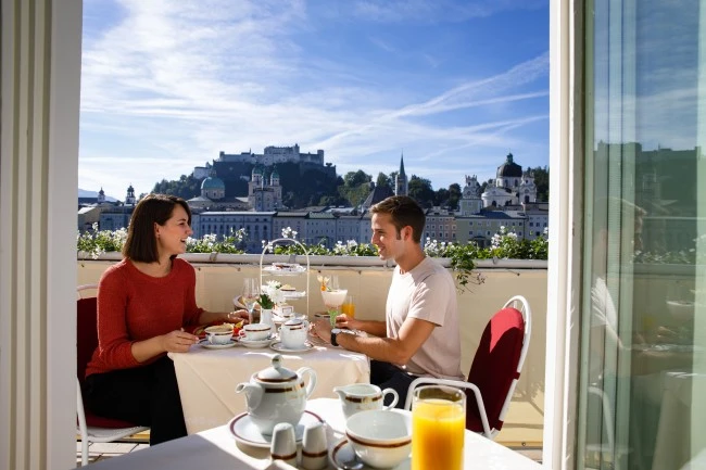 Ein Paar genießt ein Frühstück auf einer sonnigen Hotelzimmer-Terrasse mit Blick über Salzburg. Auf dem Tisch stehen Tee, Kaffee, Champagner, Säfte, Croissants und weitere Frühstücksleckereien. Im Hintergrund sind der Salzburger Dom und die Festung Hohensalzburg zu sehen.