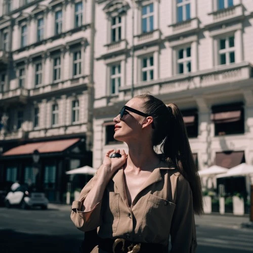 Eine Frau mit Sonnenbrille und beigem Hemdblusenkleid steht vor dem Hotel Sacher Wien und blickt in die Sonne. Im Hintergrund ist die historische Fassade des Hotels mit Markisen, Flaggen und Caféterrasse zu sehen.