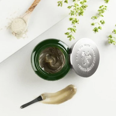 Geöffneter Tiegel mit Creme, daneben ein Spatel mit aufgetragener Produktprobe, ein Löffel mit Pulver und grüne Blütenzweige