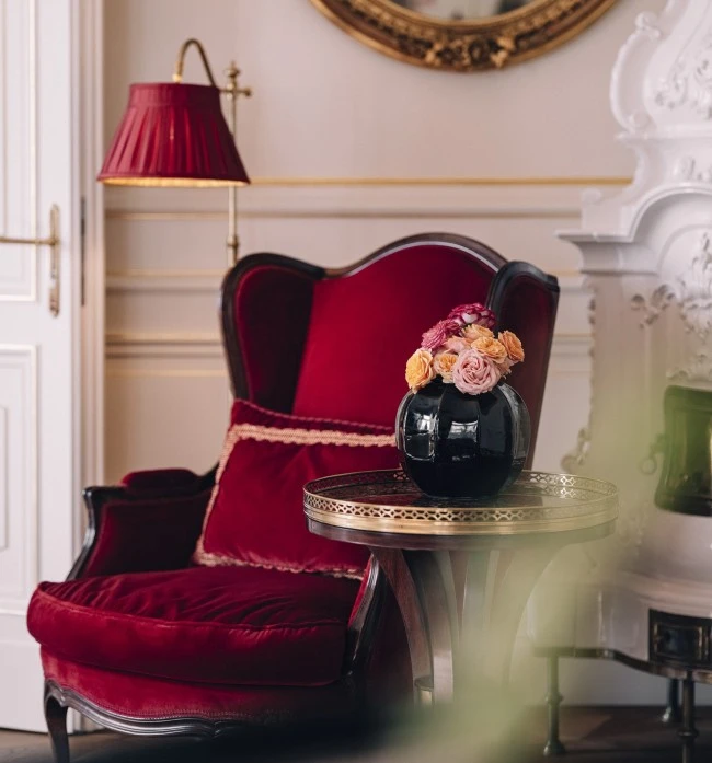 Ein Blick in die Wiener Philharmoniker Suite: Ein samtroter Ohrensessel mit passendem Kissen steht neben einem kleinen Beistelltisch mit goldener Umrandung und schwarzer Vase mit frischen Blumen. Dahinter ein traditioneller weißer Kachelofen und ein vergoldeter Spiegel.