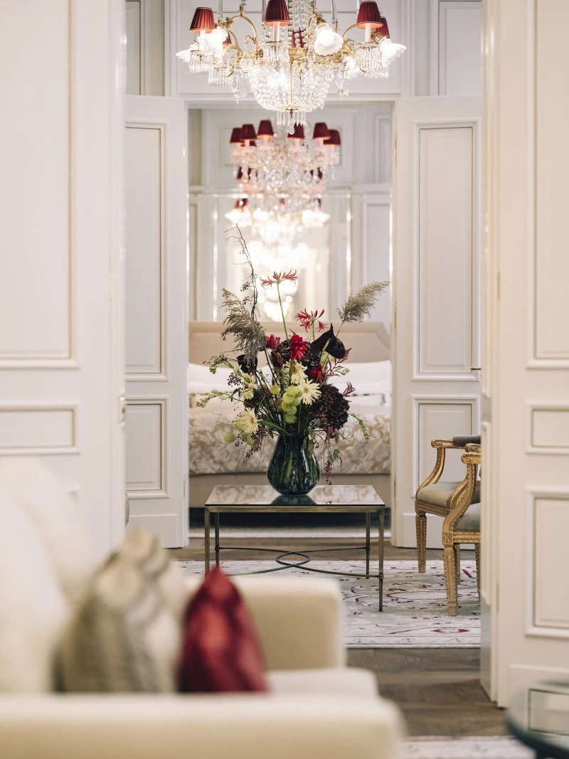 Das Bild zeigt den Durchblick vom Wohnbereich der Madame Butterfly Suite ins Schlafzimmer. Im Zentrum steht ein Glastisch mit einem Blumenarrangement, dahinter ist das Bett sichtbar. Ein Kristalllüster und ein goldener Sessel runden die elegante Szene ab.