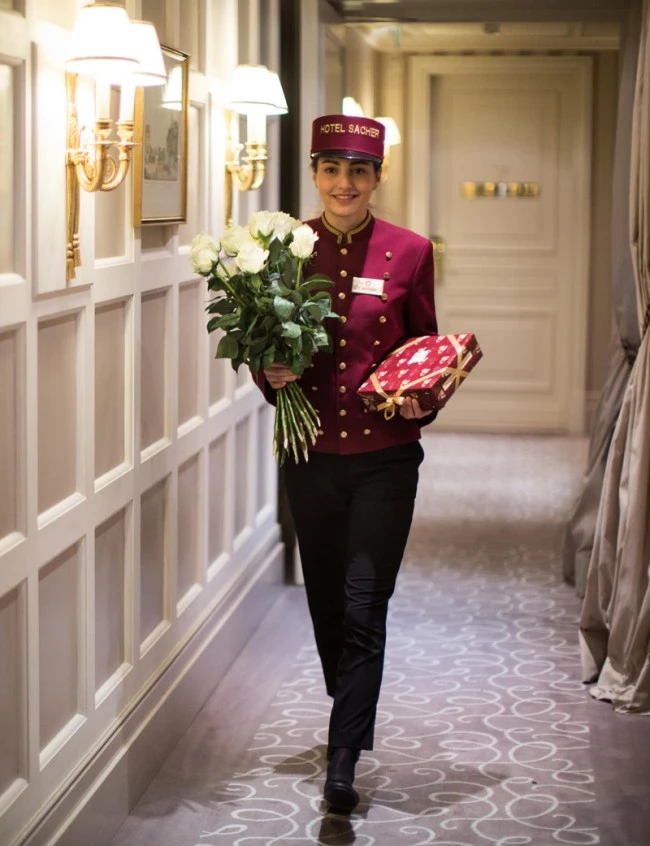 Pagin in burgunderroter Uniform bringt weiße Rosen und ein Geschenk durch einen elegant beleuchteten Hotelflur.