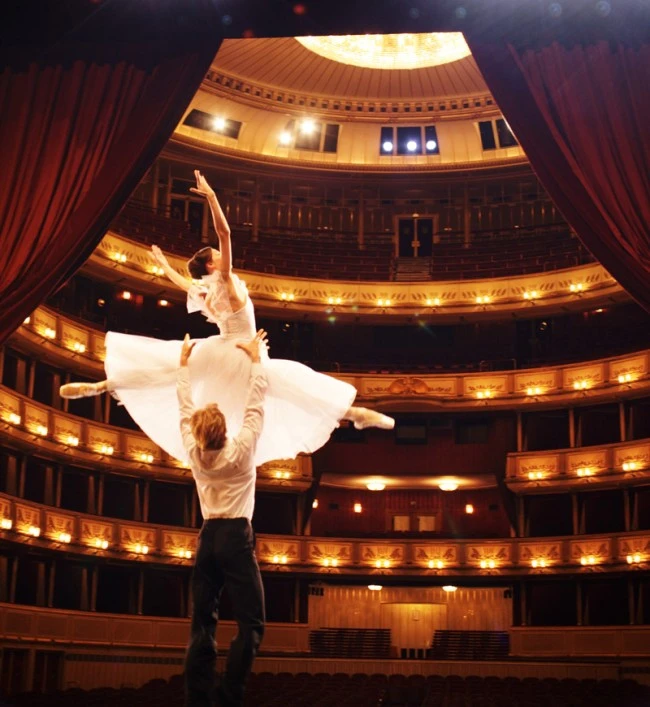 Ballettpaar auf großer Opernbühne, der Tänzer hebt die Ballerina vor majestätischem Theatersaal mit goldenen Logen und rotem Vorhang.