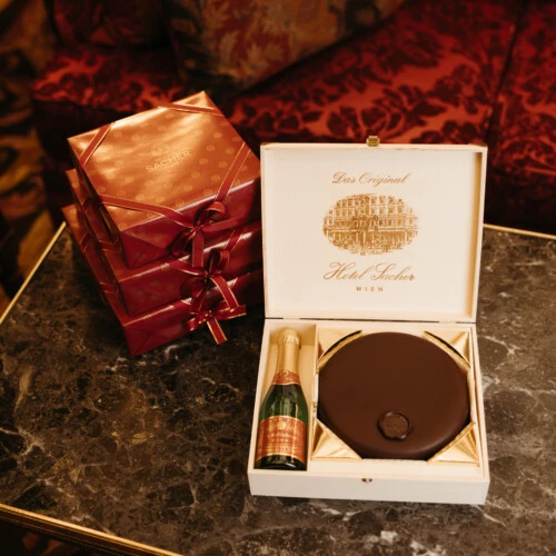 Eine geöffnete Holzkassette mit Original Sacher-Torte und einer kleinen Flasche Sekt, daneben mehrere rot-goldene Geschenkboxen, arrangiert auf einem Marmortisch.