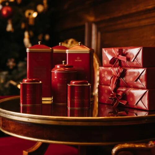 Mehrere rot-goldene Sacher-Dosen und Geschenkschachteln auf einem runden Holztisch vor einem Weihnachtsbaum.