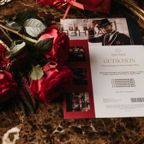 Ein Hotel Sacher Gutschein für eine Übernachtung, neben einem Strauß roter Rosen, auf einem Marmortisch platziert. Im Hintergrund eine rote Geschenkbox mit Schleife.