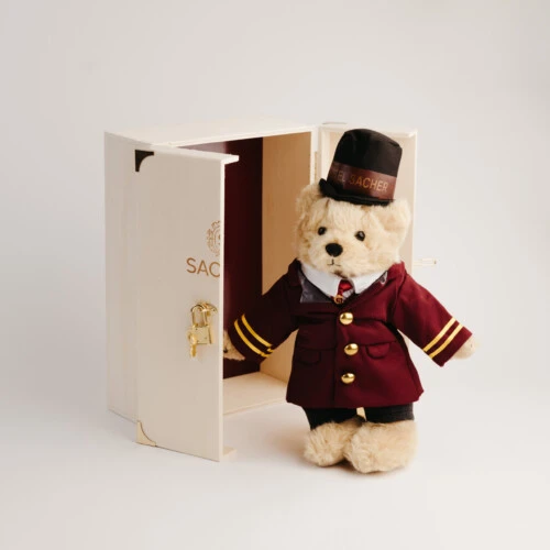 Ein Teddybär im Portier-Outfit des Hotel Sacher, mit rotem Mantel, goldenen Knöpfen und Zylinder, präsentiert vor einer offenen Holzbox mit Sacher-Logo.