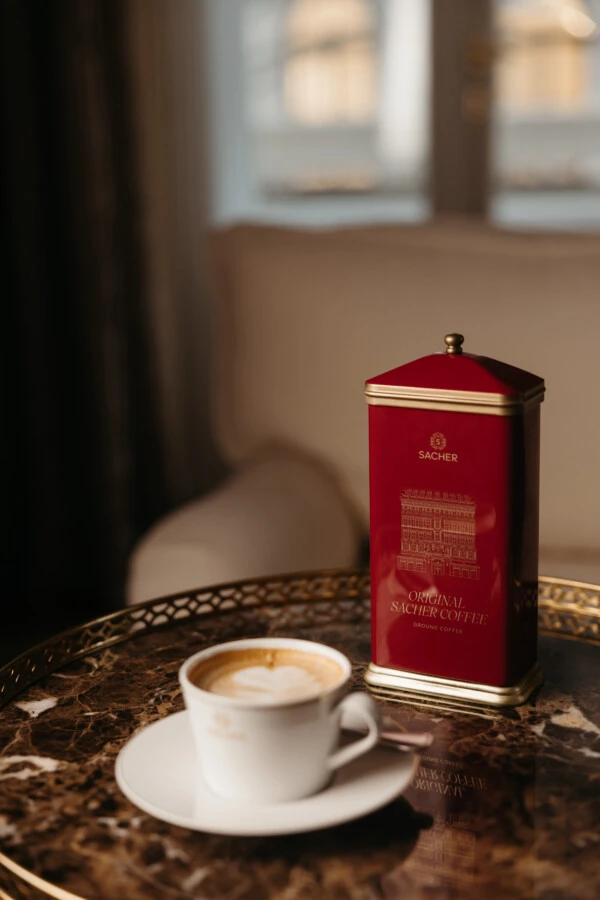 Eine Tasse Cappuccino mit Latte Art neben einer Dose Original Sacher Kaffee auf einem Marmortisch.