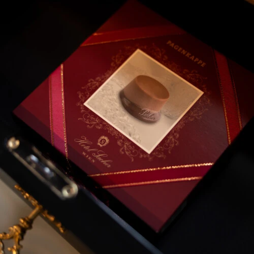 Nahaufnahme einer Geschenkbox (mit Pagenkappen-Pralinen) mit der Aufschrift „Hotel Sacher Wien“ und dem Bild einer Pagenkappe auf dem Deckel.