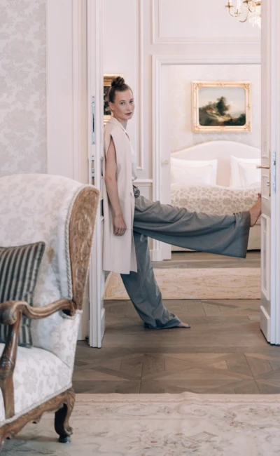 Frau in heller Weste und grauer Hose lehnt barfuß mit einem Bein an einem Türrahmen in einem elegant eingerichteten Zimmer.