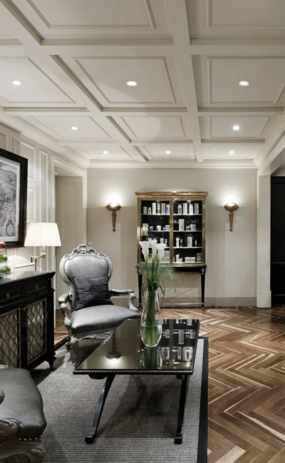 Eleganter Spa-Wartebereich mit klassischen Sesseln, einem Glastisch, Vitrine mit Kosmetikprodukten und Holzparkett im Fischgrätmuster.